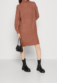 Abito a collo alto lavorato a maglia di colore ruggine, caratterizzato da maniche lunghe, texture a coste e spacchi laterali, abbinato a stivali neri chunky e una borsa.