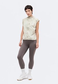 Γυναίκα όρθια, φοράει αμάνικο ανοιχτό μπεζ γιλέκο με φερμουάρ, γκρι εφαρμοστά κολάν, λευκές κάλτσες και λευκά αθλητικά παπούτσια.