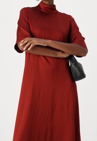 Vestido rojo de cuello alto con mangas cortas, hecho de tela suave. El modelo sostiene un pequeño bolso negro. Silueta simple y recta.