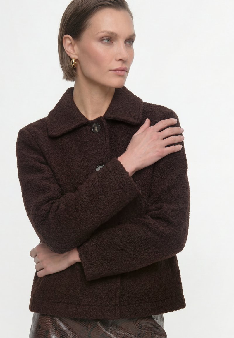Woman wearing dark brown textured jacket and gold hoop earrings, with arms crossed and looking to the side against a white background.
