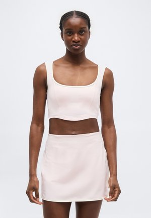 Mujer con una camiseta corta sin mangas de color beige claro y una falda de talle alto a juego, posando frente a un fondo blanco liso.