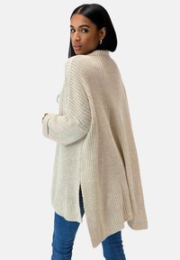 Vrouw met steil zwart haar, gekleed in een oversized beige gebreide trui en blauwe jeans, die over haar schouder kijkt tegen een witte achtergrond.