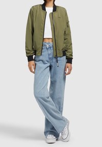 Personne portant une veste bomber vert olive, un haut court blanc, un jean large bleu clair et des baskets blanches, debout les jambes croisées.