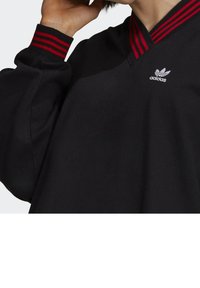 Mustat collegepaita V-päällä ja punaisilla raidallisilla yksityiskohdilla. Vasemmalla puolella kauluksen lähellä valkoinen Adidas-logo. Pehmeä rakenne.