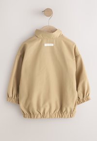 Veste beige pour enfant avec poignets et taille élastiques, suspendue à un cintre en bois devant un mur clair uni.