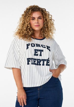 Kobieta o kręconych blond włosach stoi z jedną ręką w kieszeni, ubrana w białą koszulę w cienkie paski z napisem "FORCE ET FIERTÉ" i niebieskie dżinsy.