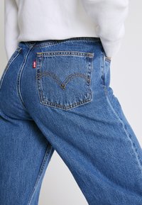Blå denimjeans med hög midja och raka ben. Funktioner inkluderar en röd etikett på fickan och karakteristisk bågformad sömnad på bakfickorna.