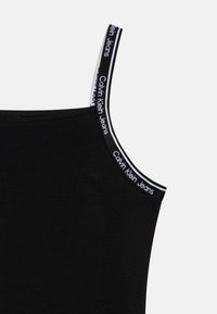 Czarny top na ramiączkach z cienkimi paskami, z białym i szarym logo "Calvin Klein Jeans" na ramieniu. Miękka, gładka tekstura materiału.