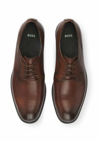 Scarpe in pelle marrone con punta arrotondata e design con lacci. Texture liscia con un leggero luccichio e suole in gomma nera. Logo del marchio visibile all'interno.