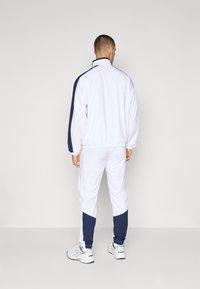 Fehér sportos melegítő egy navy kiegészítőkkel, magas gallérral, rugalmas mandzsettákkal és szűkített nadrággal. Álló pózban viseli, hátulról.