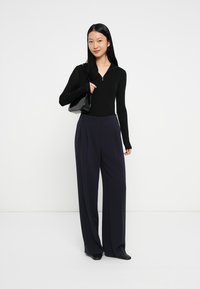 Schwarzes Langarmshirt mit Reißverschluss, kombiniert mit hochtaillierten, weiten blauen Hosen und einer eleganten schwarzen Handtasche, vor einem schlichten Hintergrund.