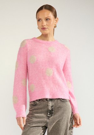 Femme portant un pull rose duveteux avec des pois beige et un jean taille haute gris sur un fond uni.