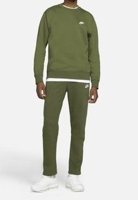 Person klädd i en olivgrön Nike-tröja och matchande joggers med vita sneakers mot en enhetlig vit bakgrund.