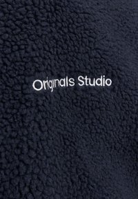 Granatowy polar z teksturowaną powierzchnią; zawiera białe haftowane napisy "Originals Studio".
