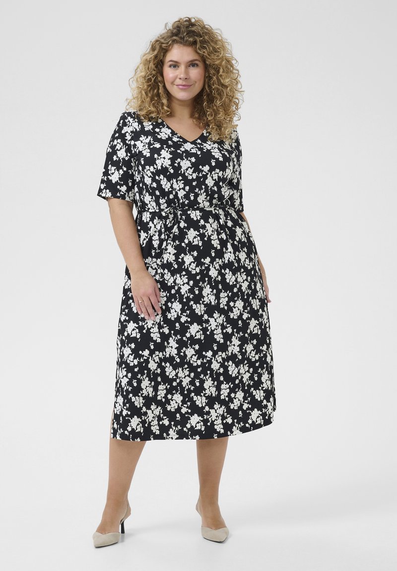 Robe noire à fleurs avec des fleurs blanches, manches courtes, décolleté en V, taille cintrée et ourlet à la cheville. Modèle portant des talons beiges.