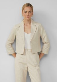 Blazer cropped beige clair en tissu texturé, avec un col à rabat, deux boutons et des manches trois-quarts, associé à un haut blanc.