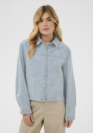 Femeie cu păr lung blond, purtând o jachetă din denim cu nasturi, albastru deschis cu model, și pantaloni bej, privindu-se înainte pe un fundal alb.