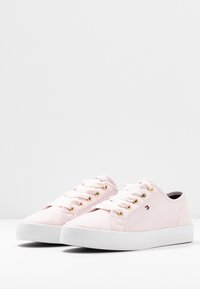 Růžové plátěné tenisky se zlatými očky a bílými gumovými podrážkami. Mají ploché tkaničky a nenápadný detail loga na straně.