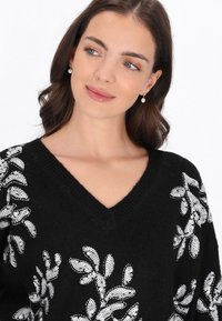 Czarny sweter dzianinowy z dekoltem w serek, zdobiony białymi cekinowymi wzorami kwiatowymi na rękawach. Tekstura wydaje się miękka, a krój jest luźny.