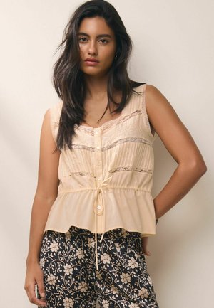 Femeie cu păr lung și închis la culoare, purtând un top bej fără mâneci, cu nasturi și detalii din dantelă și pantaloni negri cu model floral.