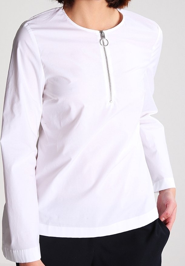 Camicia bianca a maniche lunghe con scollo rotondo e una cerniera frontale con tirante circolare. Tessuto liscio con una vestibilità aderente. Pantaloni neri non visibili.