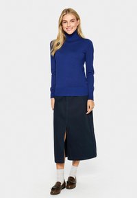 Mėlyna turtlenecko tipo nėrinių megztinis su ilgomis rankovėmis, derinamas su tamsiomis sijonais, kurie turi priekinę skeltą. Modelis vilki rudus loafer’ius ir pilkas kojines.