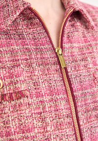 Vaaleanpunainen tweed-takki, jossa on teksturoitu kuvio, kultainen vetoketju ja viininpunaisia yksityiskohtia. Takissa on kaulus ja vetoketjullinen etuaukko.