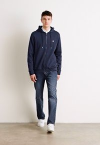 Marineblauwe hoodie met rits en trekkoorden, gedragen over een witte blouse, gecombineerd met donkere spijkerbroek en witte sneakers; zachte stofstructuur.