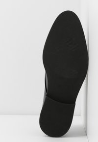 Semelle de chaussure formelle noire avec bout pointu et talon texturé, présentée sur un fond blanc d'angle.