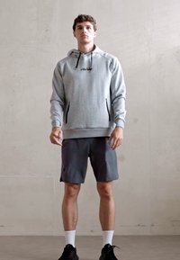 Pelēks hūdis ar melniem akcentiem, ar priekšējo logo, saskaņots ar tumšiem šortiem. Modelis valkā melnus sporta apavus un baltas apakšbikšu zeķes.