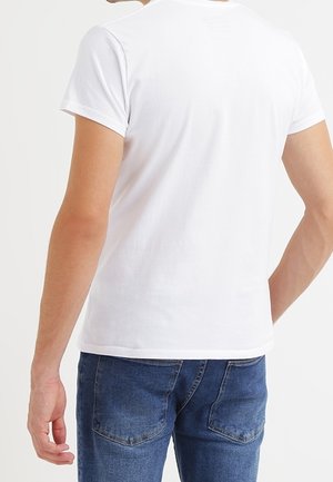 Mand iført hvid, ensfarvet T-shirt med korte ærmer og blå jeans, set fra ryggen mod en ensartet lys baggrund.