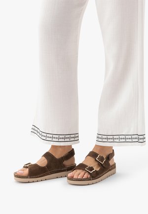 Stopy w brązowych sandałach zamszowych z dwoma paskami zapinanymi na klamry, zestawione z białymi spodniami o szerokich nogawkach z czarnymi haftowanymi brzegami.