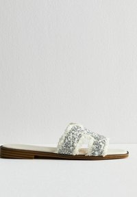 Vita sandaler i vitt med dekorativa silverdetaljer, fransar på remmarna och en platt brun träsoff.