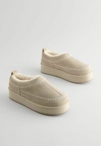 Bottines à plateforme beiges à enfiler avec des coutures blanches et une doublure en peluche, présentées en biais sur un fond blanc.