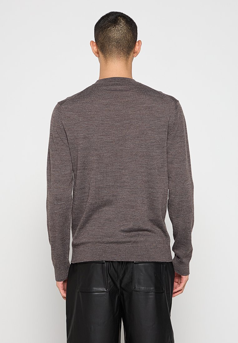 Hombre vestido con un suéter marrón de manga larga y pantalones de cuero negros, de pie de espaldas contra un fondo blanco liso.