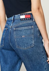 Blugi din denim albastru cu talie înaltă, având o etichetă cu logo-ul Tommy Jeans pe spate și două buzunare posterioare cu cusături vizibile.