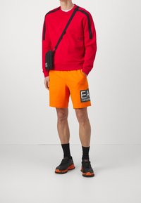 Sudadera roja con detalles en negro, pantalones cortos naranjas con un logo, calcetas negras y zapatillas negras-naranjas con suelas texturizadas.