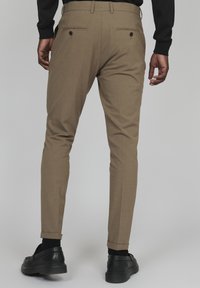 Bež hlače s slim fit krojem, z dekorativnimi zadnjimi žepi in zavihanimi hlačnicami, oblečene z črnimi čevlji in nogavicami.