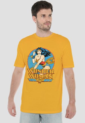 Mann trägt ein gelbes T-Shirt mit einem bunten Vintage Wonder Woman-Grafik und Text auf der Vorderseite, steht vor einem schlichten Hintergrund.