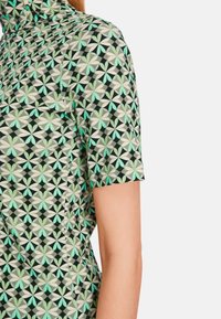 Kurzarmhemd mit schwarzem Grund und geometrischen Mustern in Lindgrün, Mint und Creme. Weicher Stoff mit tailliertem Design.