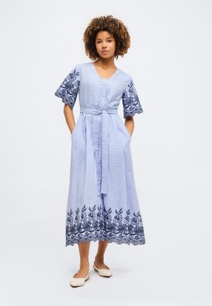 Γυναίκα στέκεται φορώντας ένα ανοιχτό μπλε μίντι φόρεμα με ναυτικό κεντητό σχέδιο στα μανίκια και το τελείωμα, δεμένη ζώνη και κρεμ μπαλέτα.