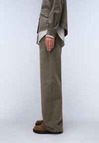 Ελαιόλαδο πράσινα βαμβακερά παντελόνια και ελαφριά γκρι ριγέ πουκάμισο, με άνετη εφαρμογή. Καφέ υποδήματα ολοκληρώνουν την εμφάνιση, με ορατές τσέπες.