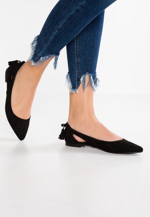 Fødder iført sorte flade sko med spidse snuder og knapper bagpå, kombineret med mørkeblå skinny jeans med frynset kant mod en hvid baggrund.
