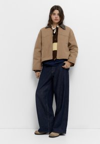 Tan cropped jakke med en brun og creme-stribet sweater, mørke brede jeans og beige ankelstøvler, med knaplukning og sidelommer.