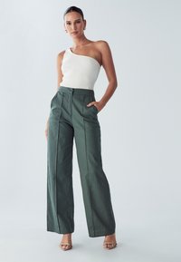 Witte geribbelde top met één schouder, gecombineerd met hooggeknoopte, wijduitlopende groene broeken met zijzakken en een geplooide voorkant. Naakte sandalen accentueren de outfit.