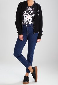Černá bomber bunda na zip přes tričko s grafickým motivem v černobílé, spárovaná s tmavě modrými skinny džíny a černými teniskami s hnědými podrážkami.