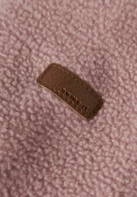 Zacht roze fleece stof met een klein bruin leren etiket dat is gestikt, met het merknaam "name it." erin geperst.