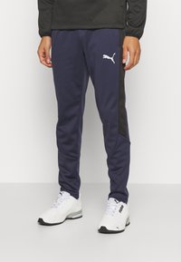 Námornícky sportovní kalhoty s texturovaným designem, černým bočním panelem a bílým logem Puma. Kombinováno s bílými sportovními botami.