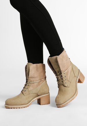 Beżowe zamszowe i skórzane botki na sznurowadła z szerokimi obcasami, noszone z czarnymi dopasowanymi spodniami na białym tle.