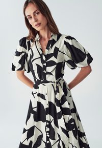 Schwarz-weißes Kleid mit floralem Muster, Knopfleiste vorne, kurzen Puffärmeln und einem Gürtel in der Taille. Glatter Stoff.